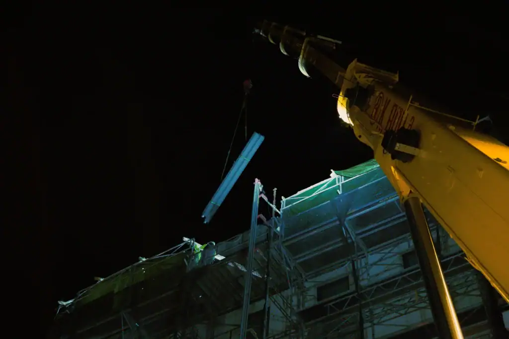 Kran løfter konstruksjonsmaterialer til byggeplass om kvelden under mørke forhold.